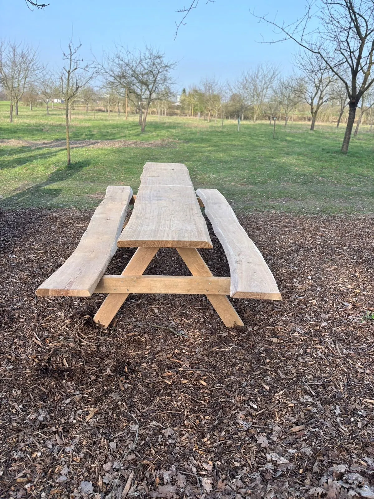 Tisch-Bank-Garnitur aus massivem Holz auf Hackschnitzeln vor einer Obstwiese.